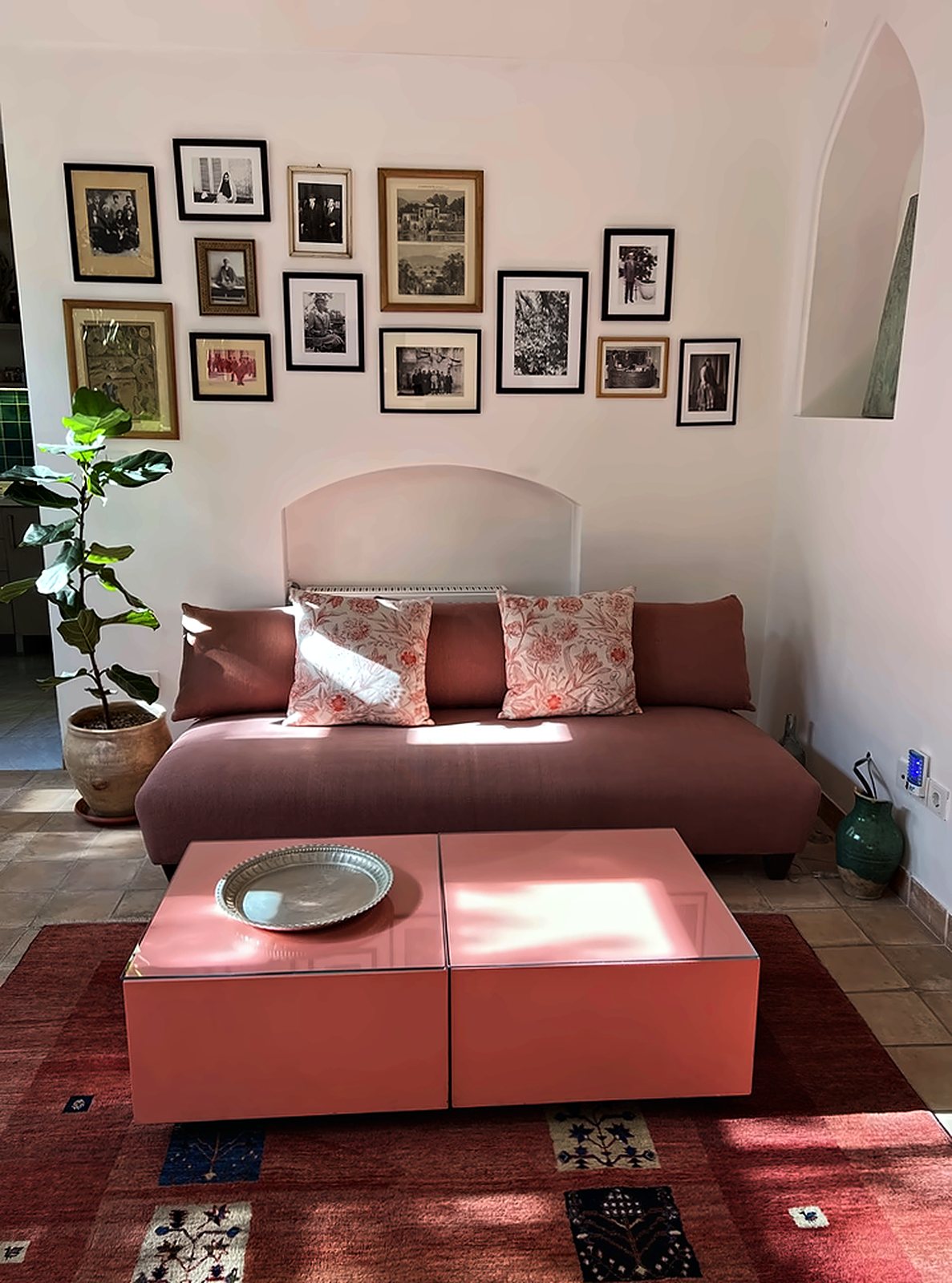 The living room: a dusty-pink sofa beneath a wall of framed family photographs, a low pink table and a red rug in the morning sun